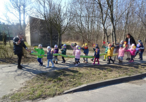 Dzieci wraz z paniami maszerują z wężem ulicami osiedla, w ręku trzymają uniesione papierowe kwiatki, śpiewają wiosenne piosenki.